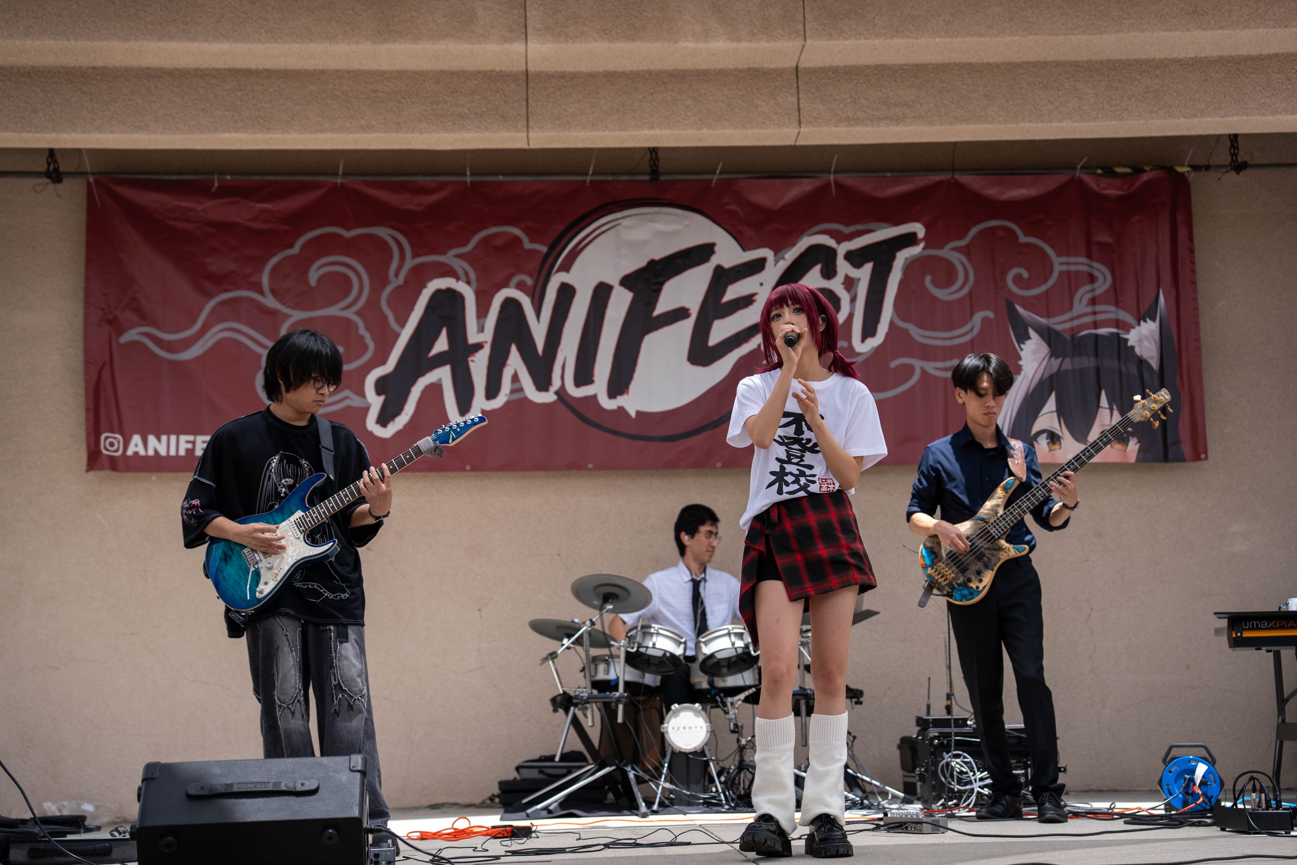 Four-piece band performing on an outdoor stage beneath a red ANIFEST banner, vocalist mid-note at the microphone
