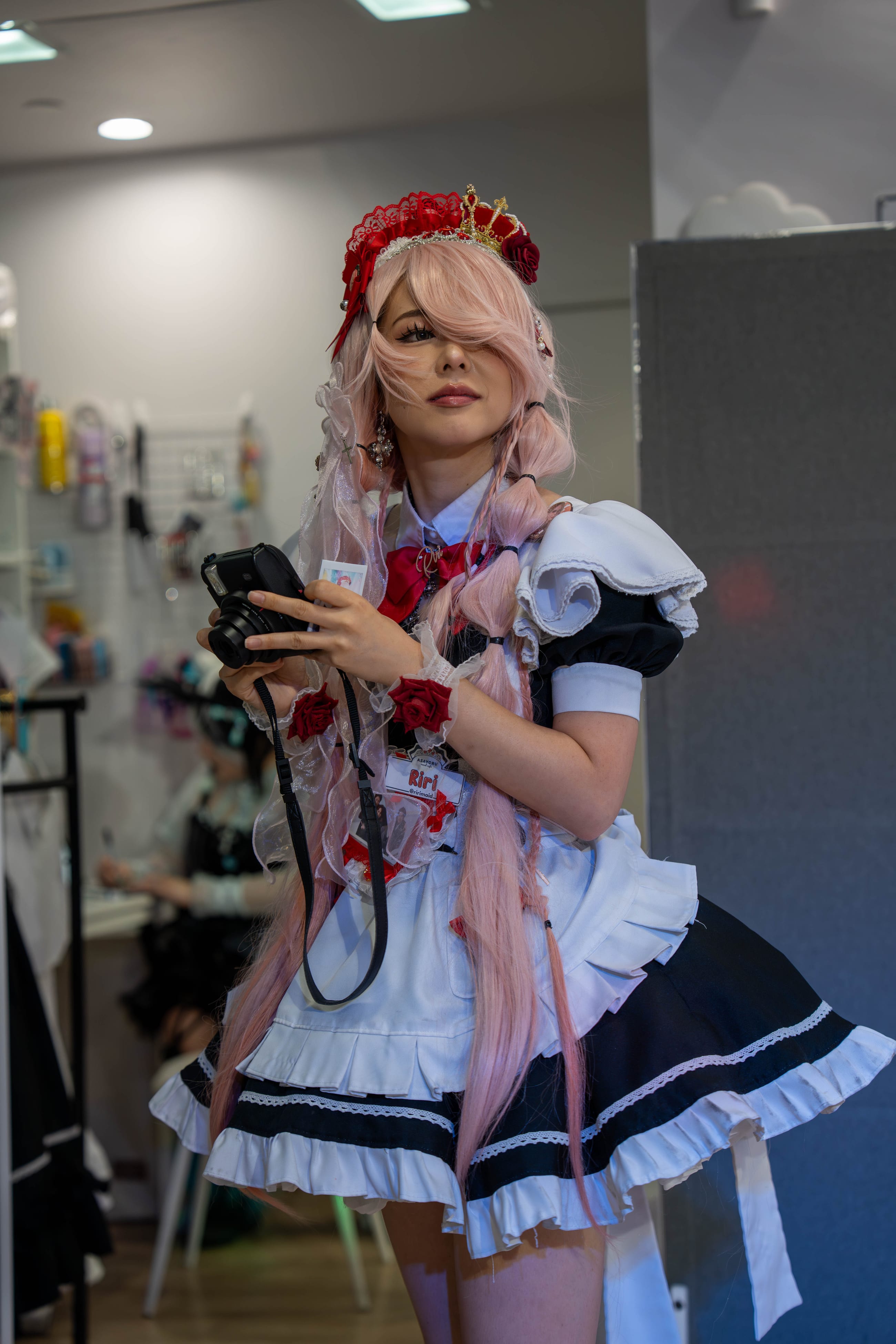 Cosplayer in a maid uniform with pink hair and a red headpiece, holding a camera, cafe interior behind