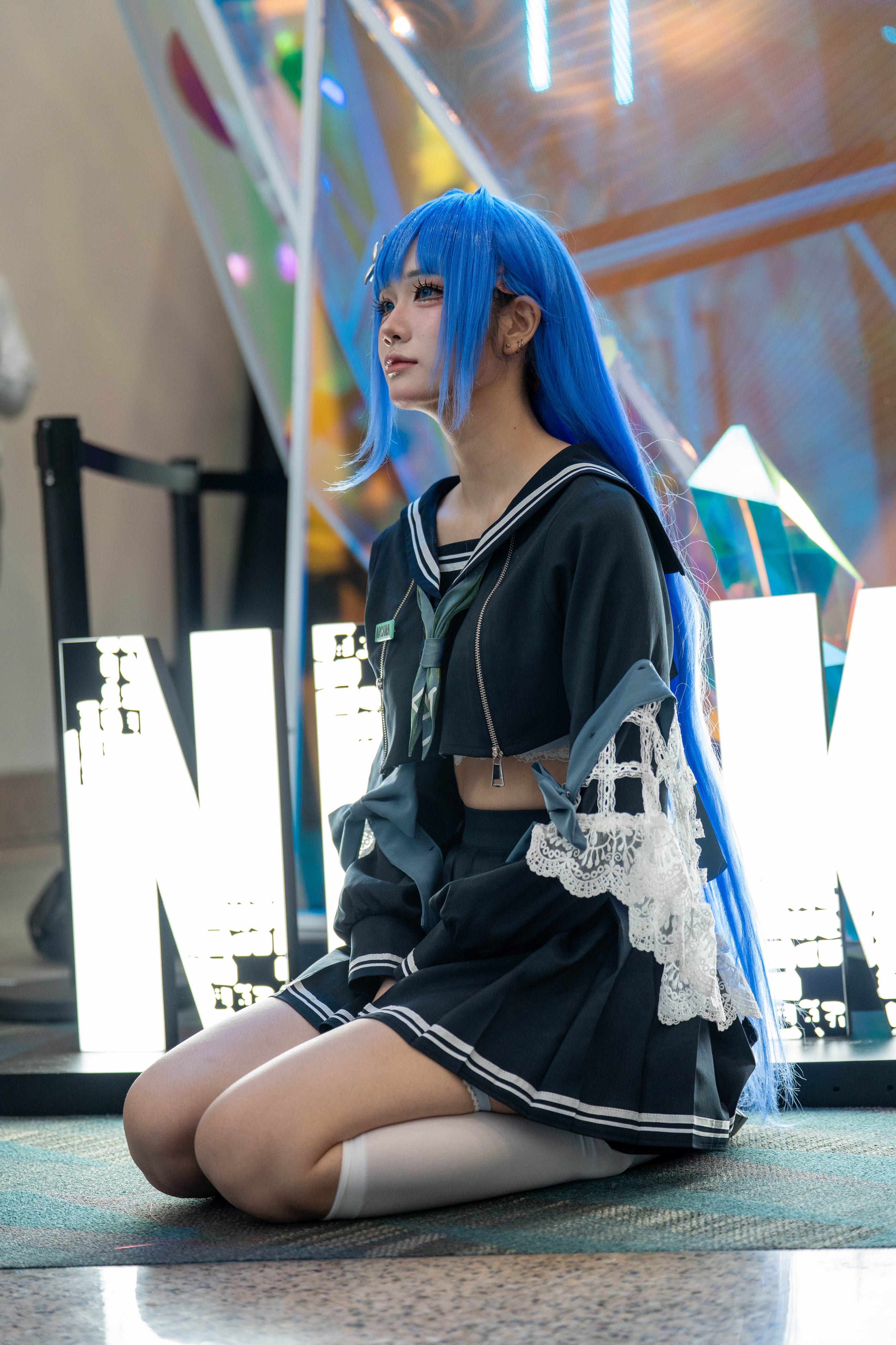 Cosplayer with long blue hair kneeling in front of illuminated letters at a brand activation