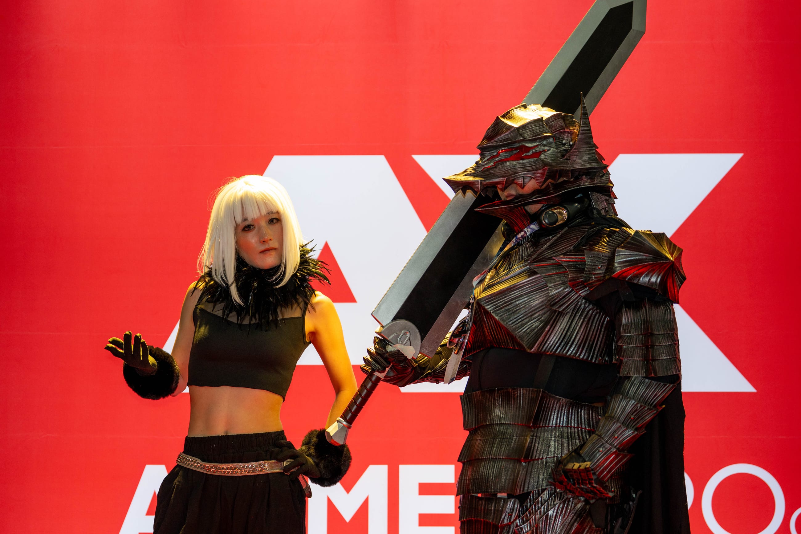 Two cosplayers in armor and a broad sword posed in front of an enormous red AX ANIME EXPO wall