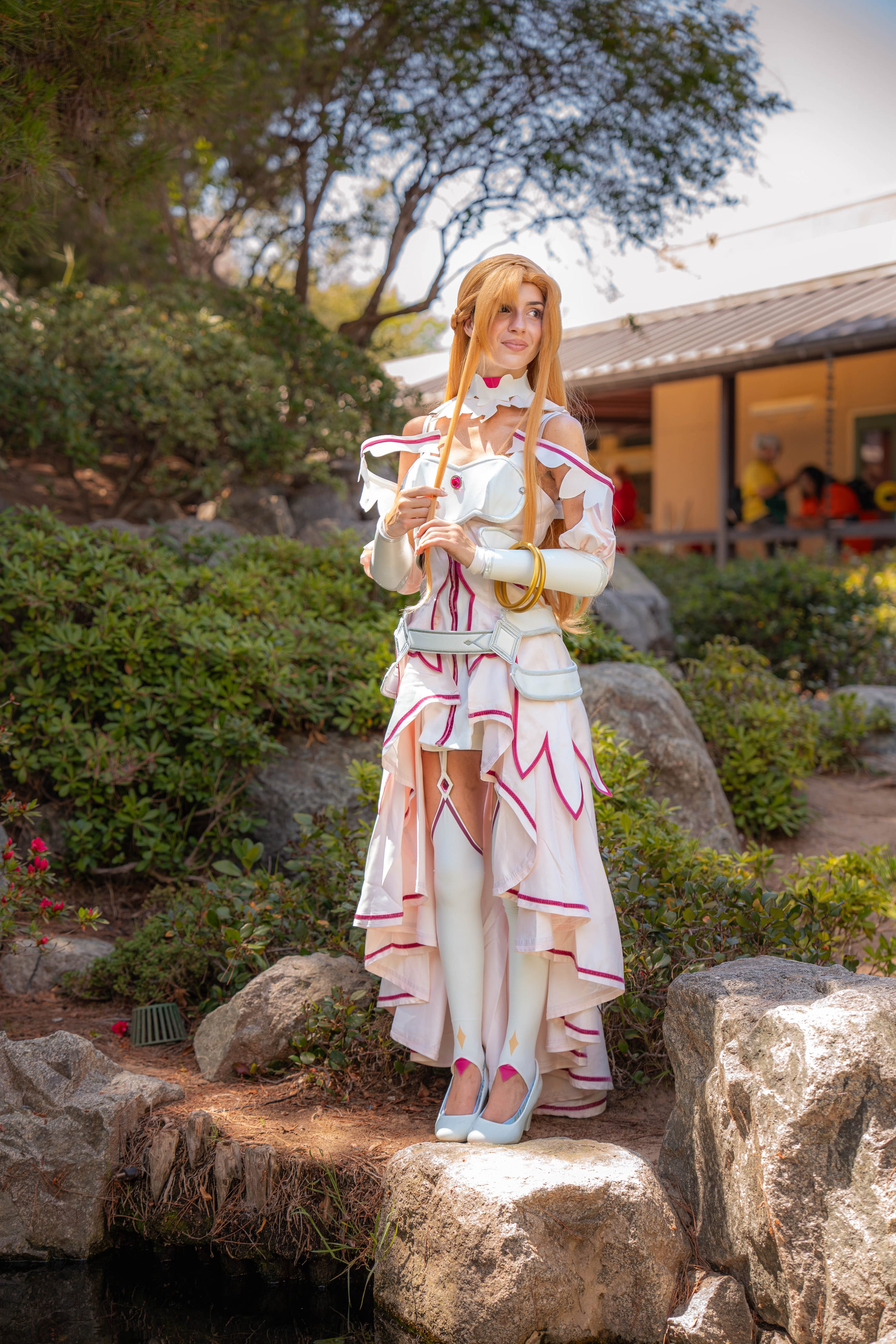 Cosplayer as Asuna standing on stone in a garden, hands clasped, looking toward camera in warm afternoon light