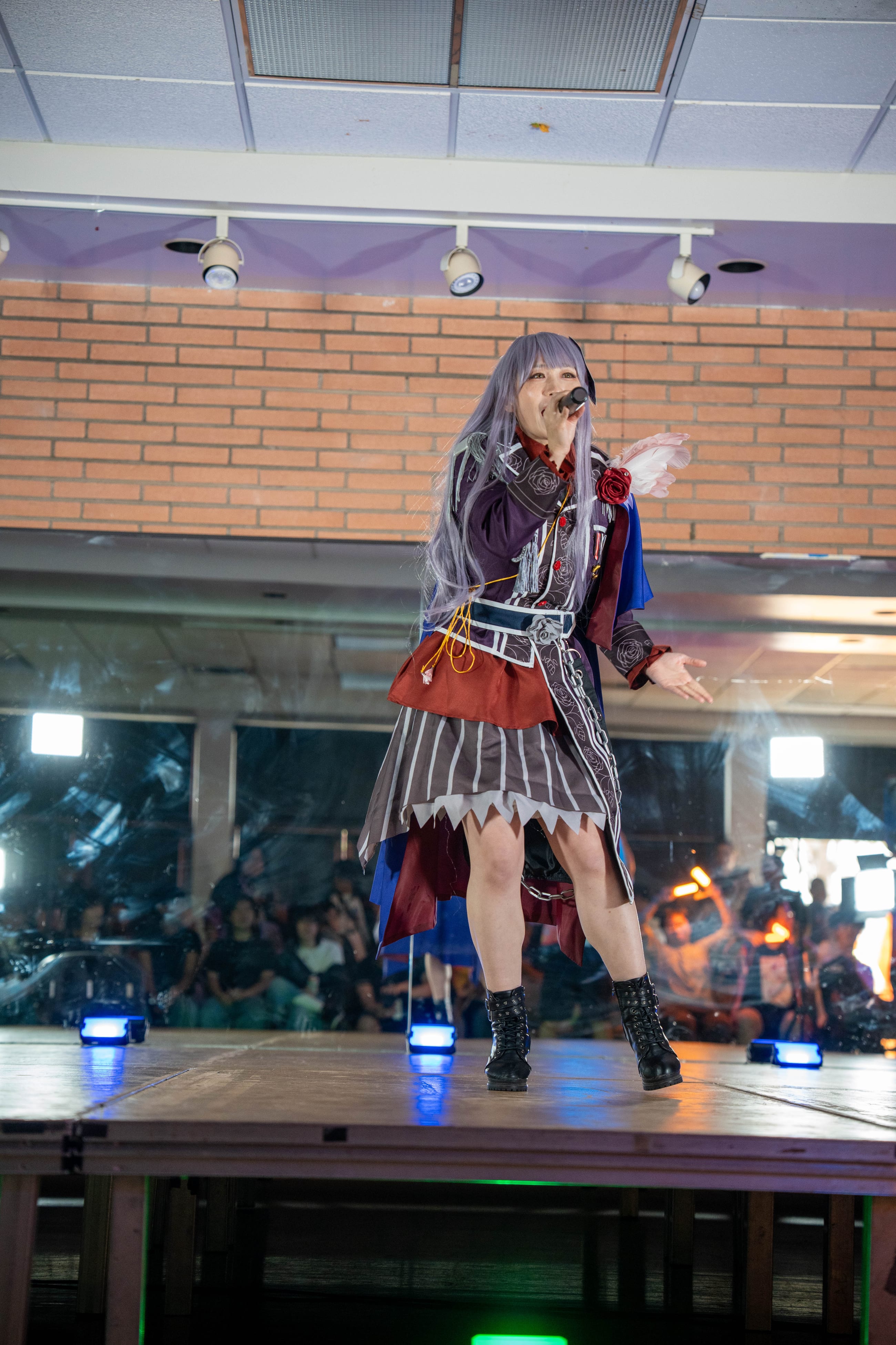 Cosplayer walking a lit runway in a pleated skirt and boots, audience seated in the dark beyond