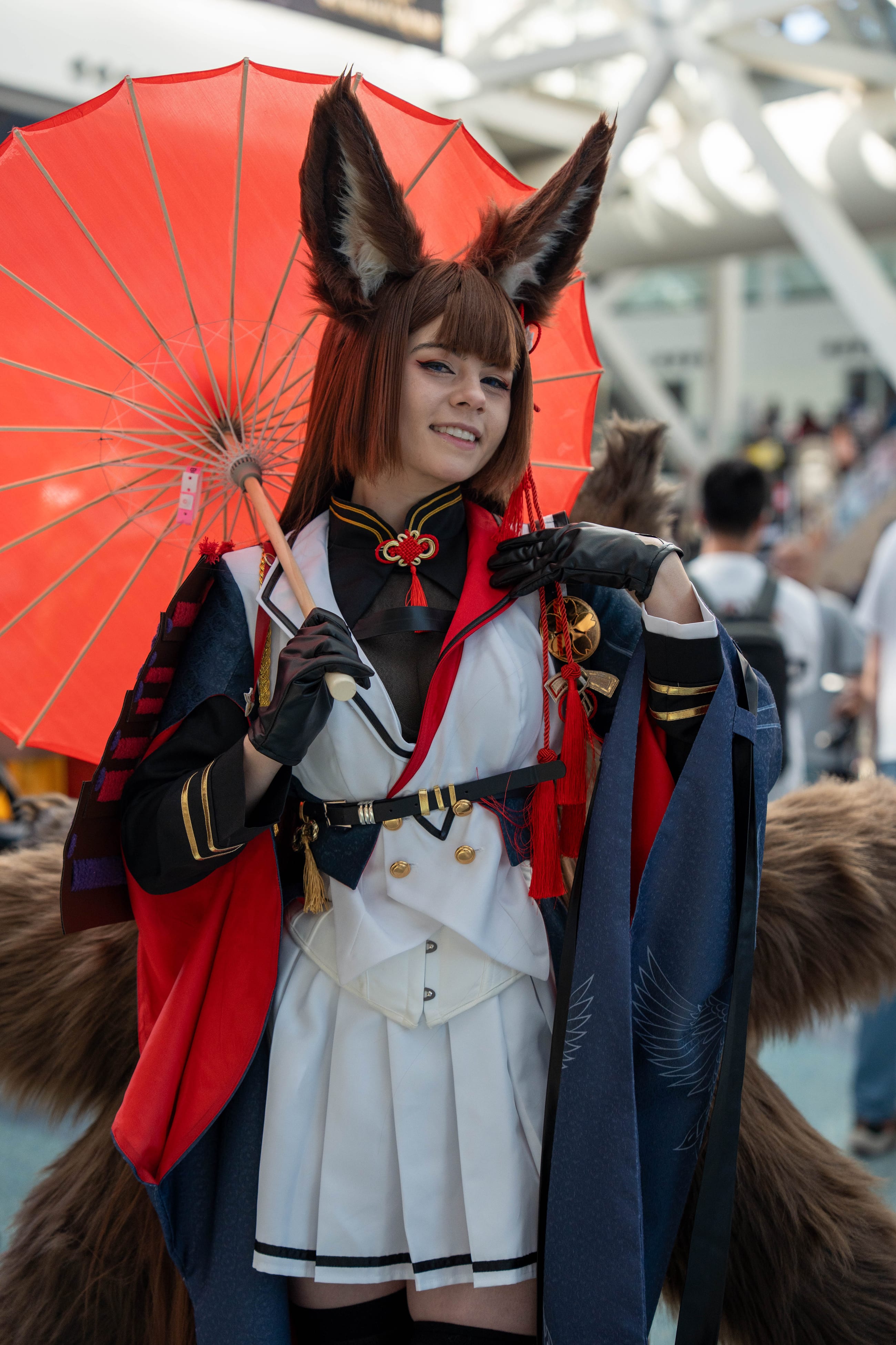 Cosplayer with fox ears and a red paper parasol, in a white and navy uniform with a fur tail