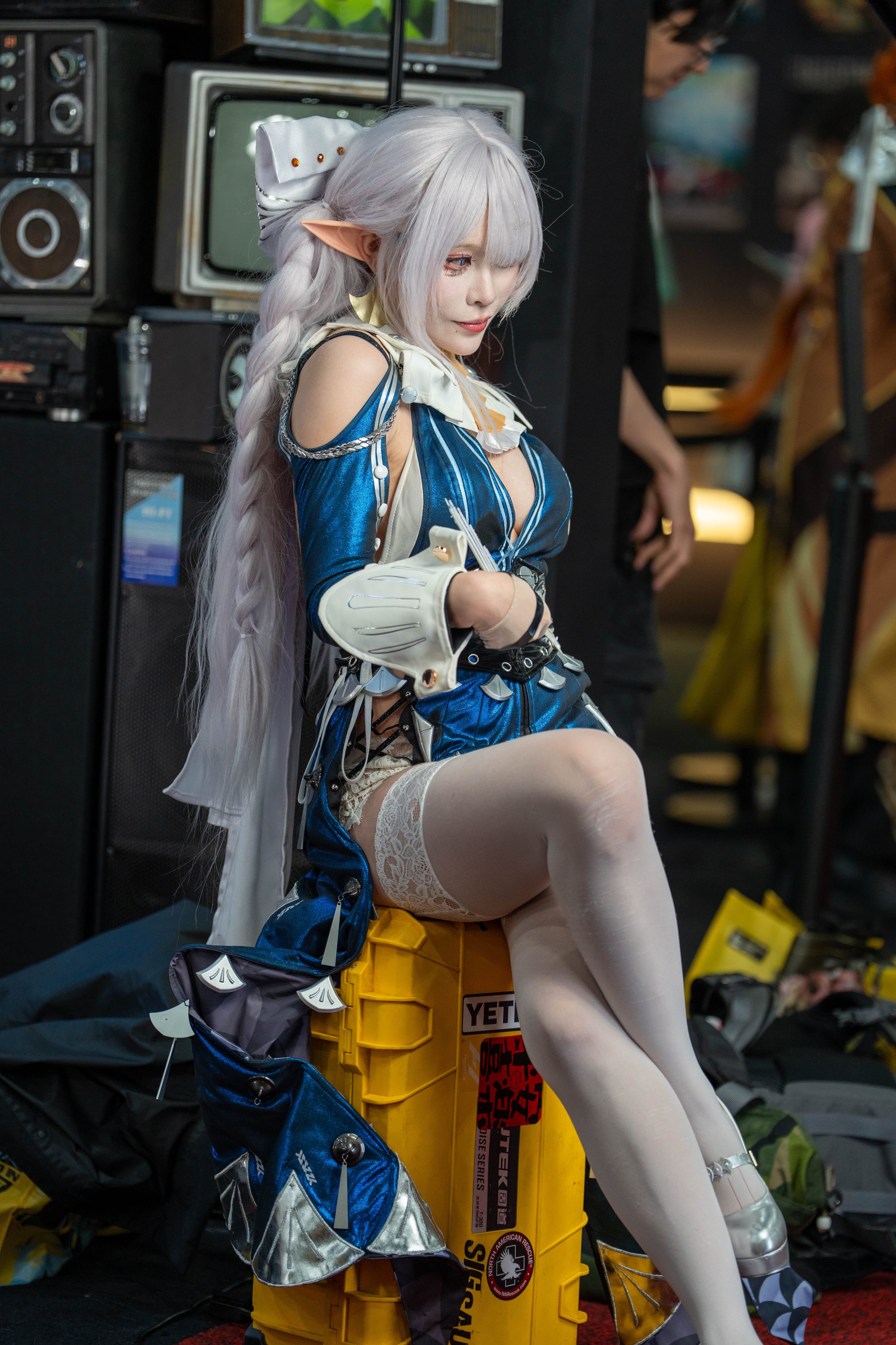 Cosplayer in blue and white armor seated on a yellow equipment case in front of a wall of stacked vintage televisions