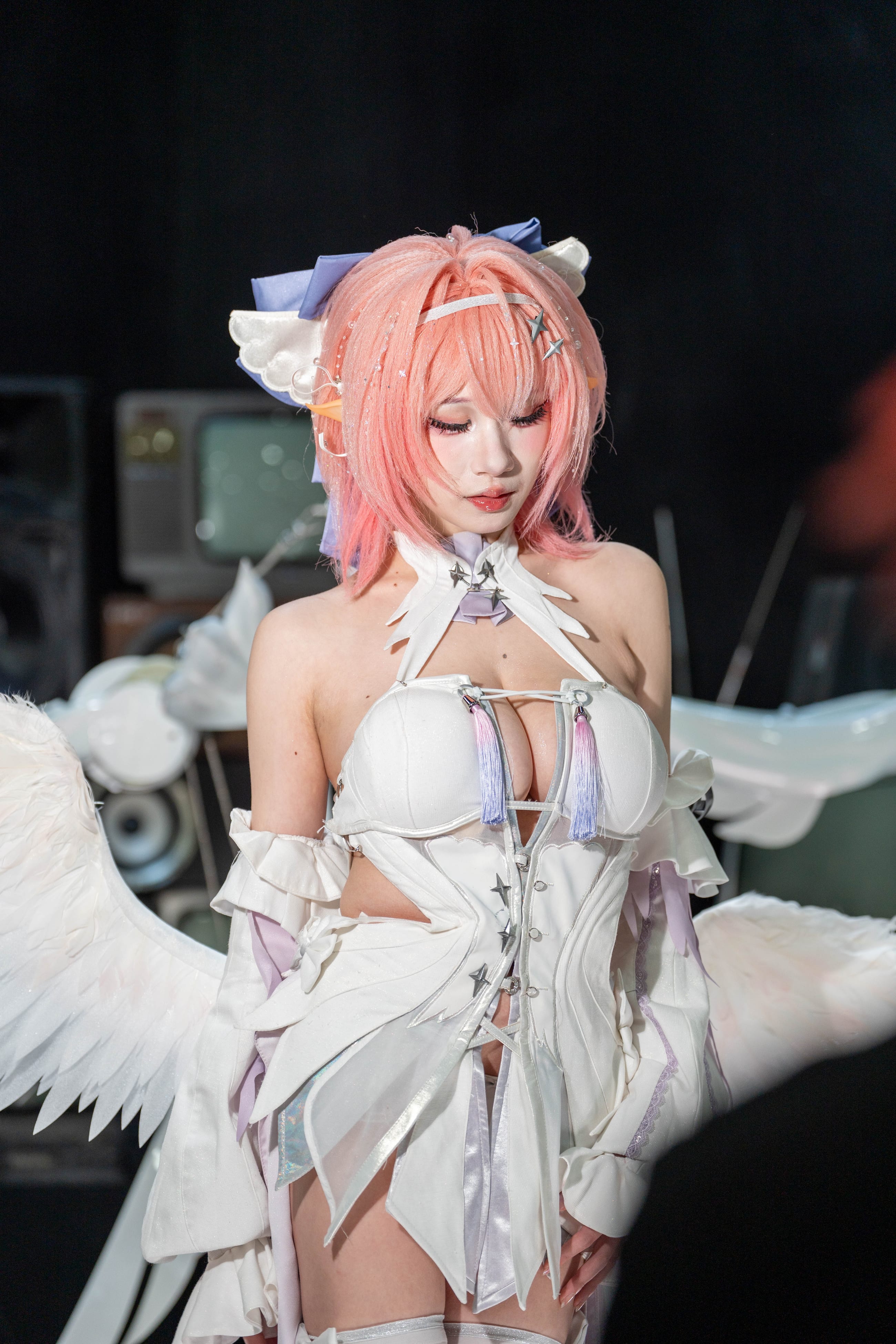 Cosplayer with pink hair and layered white feathered wings against a dark cluttered set
