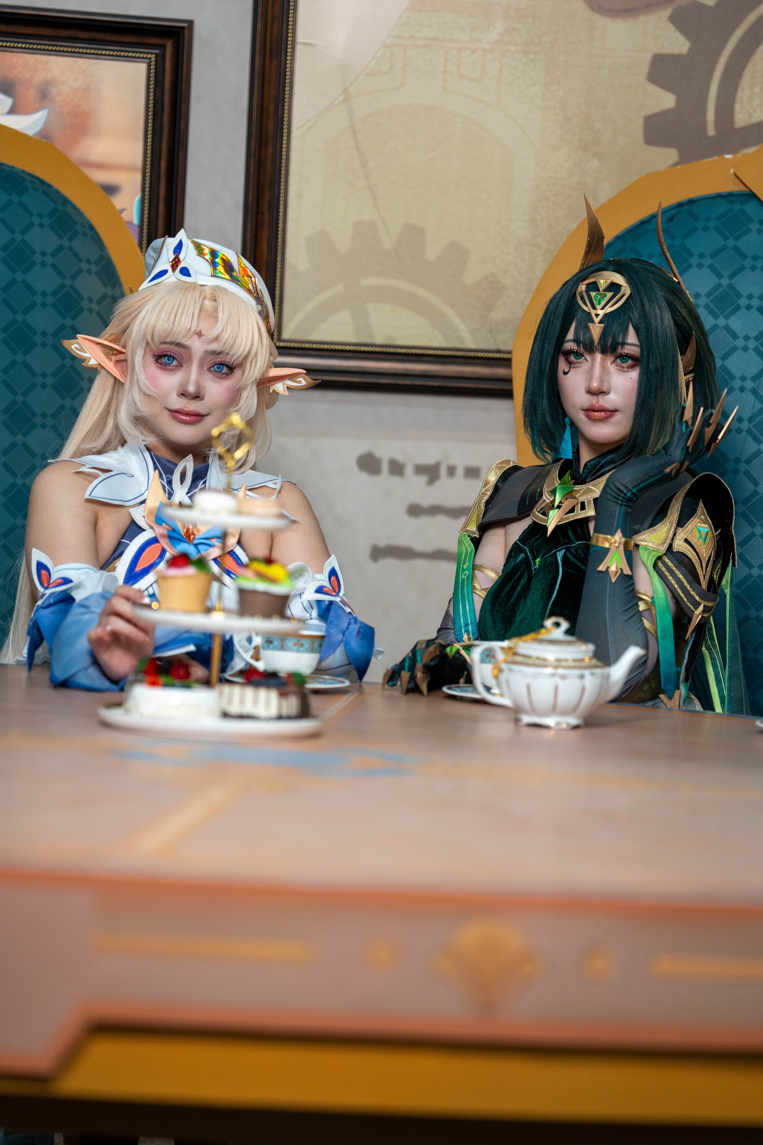 Two cosplayers seated at a themed cafe activation with a tiered stand and teapot on the table between them