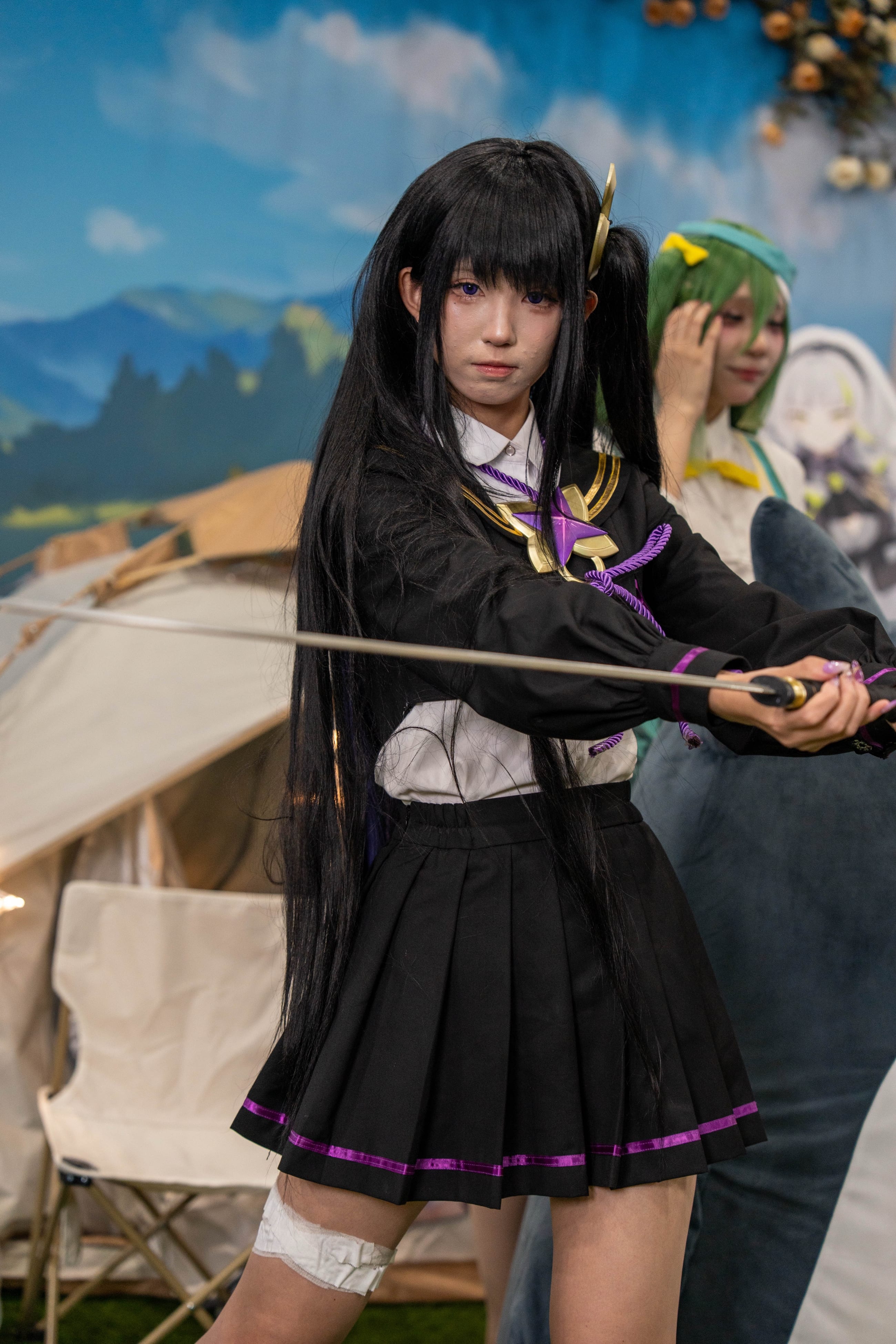 Cosplayer in a black school uniform drawing a long blade, painted sky backdrop behind her